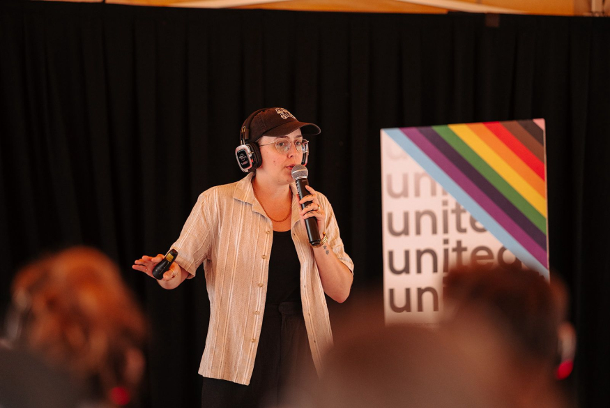 Collega Anke geeft vorming, met microfoon en regenboogvlag op achtergrond