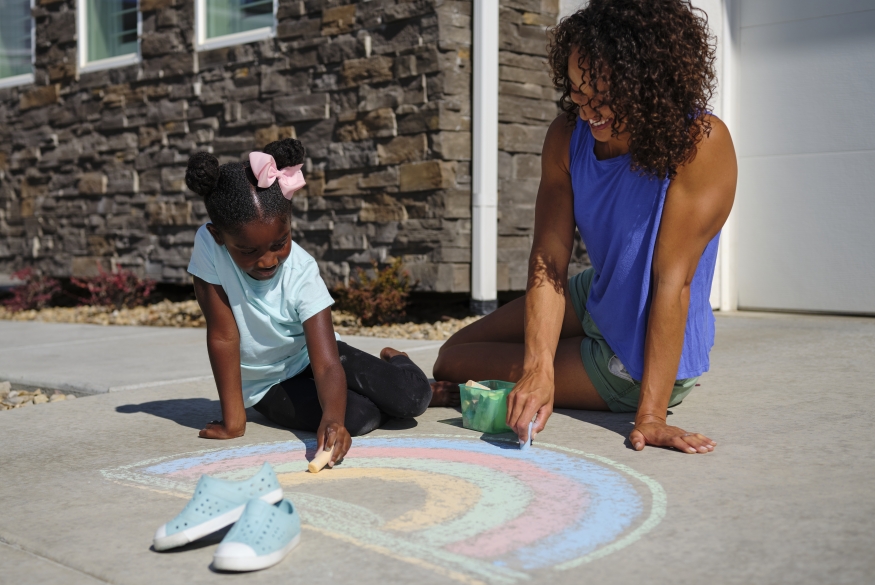 Een zwart kind en zwarte volwassene die samen al lachend een regenboog tekenen met stoepkrijt