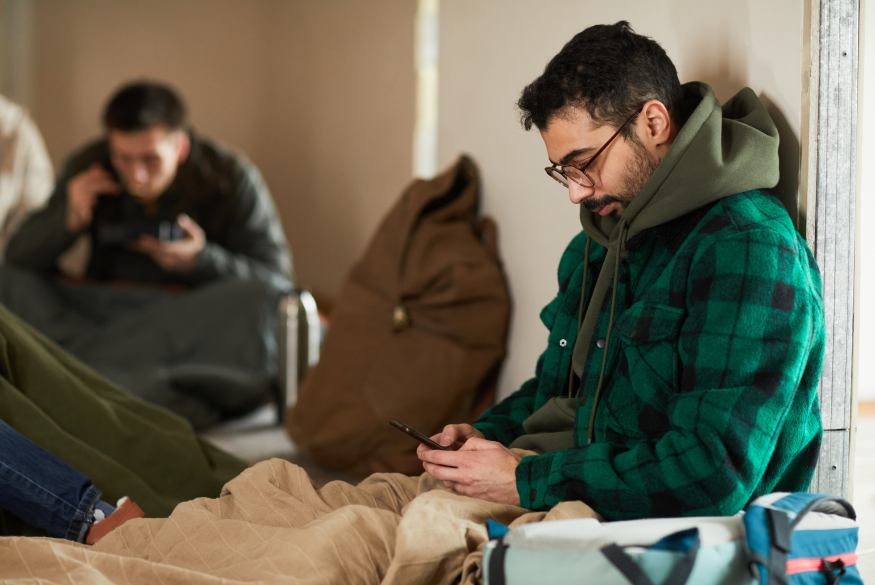 Persoon die neerzit in een asielcentrum en hun gsm vastheeft met links een persoon op de achtergrond die aan het eten is