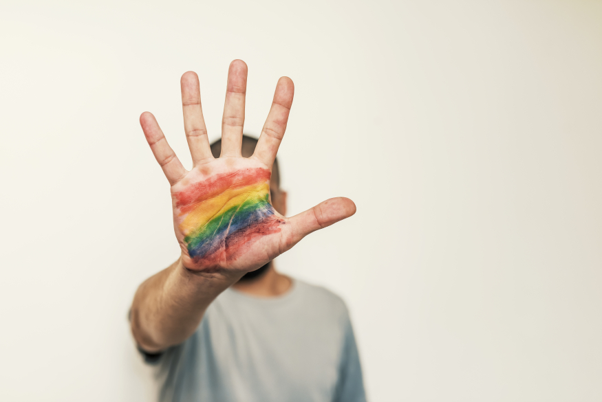 Iemand maakt het stopteken met hun hand. Op het hand staat een regenboogvlag gekleurd. 