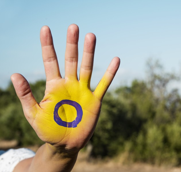 Iemand toont hun hand in de stop houding. Hun hand is geel geschilderd met een paarse cirkel in het midden, het symbool voor de intersekse vlag. 
