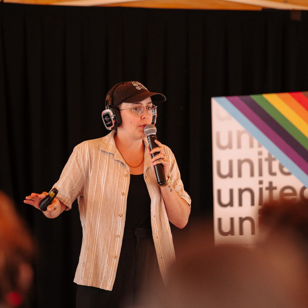 Collega Anke geeft vorming, met microfoon en regenboogvlag op achtergrond